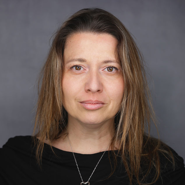 A professional headshot of a light-skinned woman with shoulder-length brown hair. She wears a black blouse and a necklace.